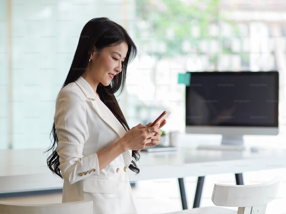 Side view of businesswoman smiling and using smartphone while standing in office room