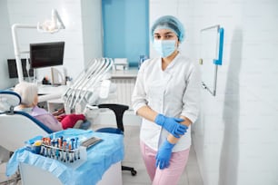 Lady in white lab coat and pink pants with disposable hat and face mask grabbing left arm with right hand