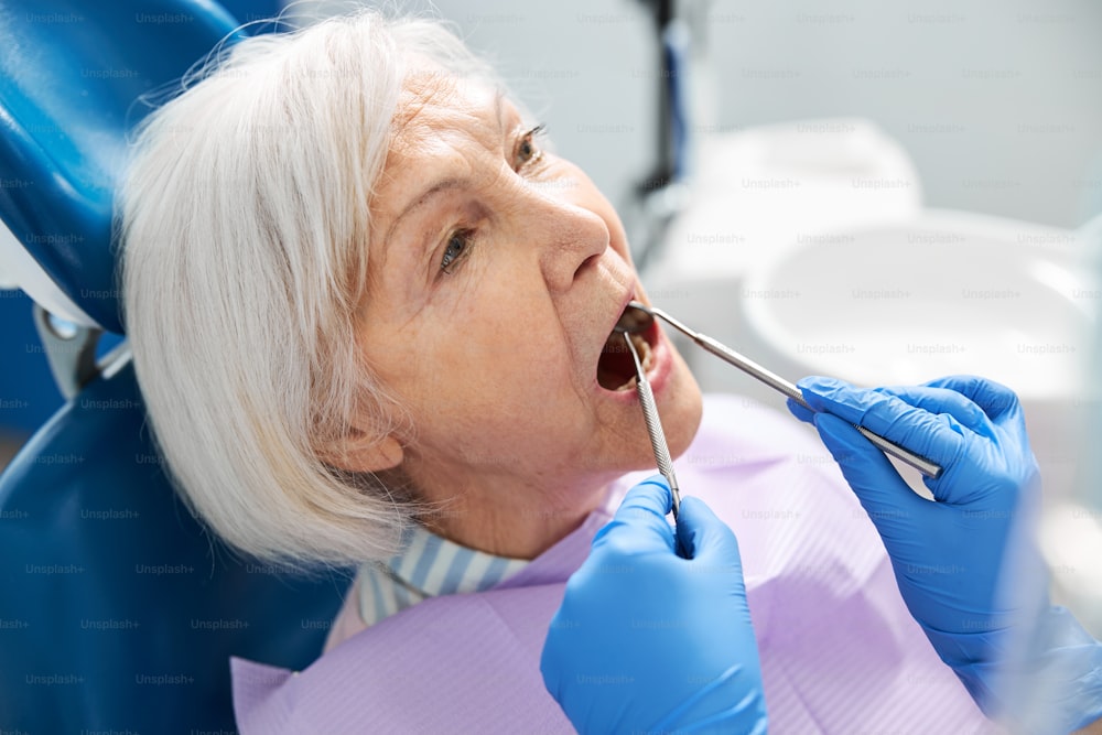 Aged female lying in dental chair with opened mouth while doctor checking her teeth with probe and mirror