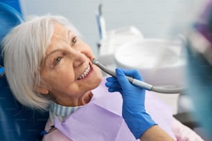 Zahnklinikarbeiter, der mit einem Handstück in der Hand an älteren erwachsenen Zähnen operiert und einen sterilen blauen Handschuh trägt