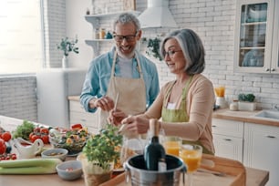 Glückliches älteres Paar in Schürzen, das ein gesundes Abendessen zubereitet und lächelt, während es Zeit zu Hause verbringt