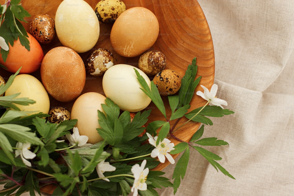 ¡Felices Pascuas! Huevos de pascua modernos en cuenco de madera con flores de primavera sobre tela de lino rústico, vista superior con espacio para texto.  Huevos teñidos al natural sobre tela gris con flores de anémona en flor. Estético