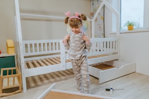 Niña de 4 años con pijama y disfraz de alas de mariposa jugando en la habitación de los niños cerca de la cama nueva.