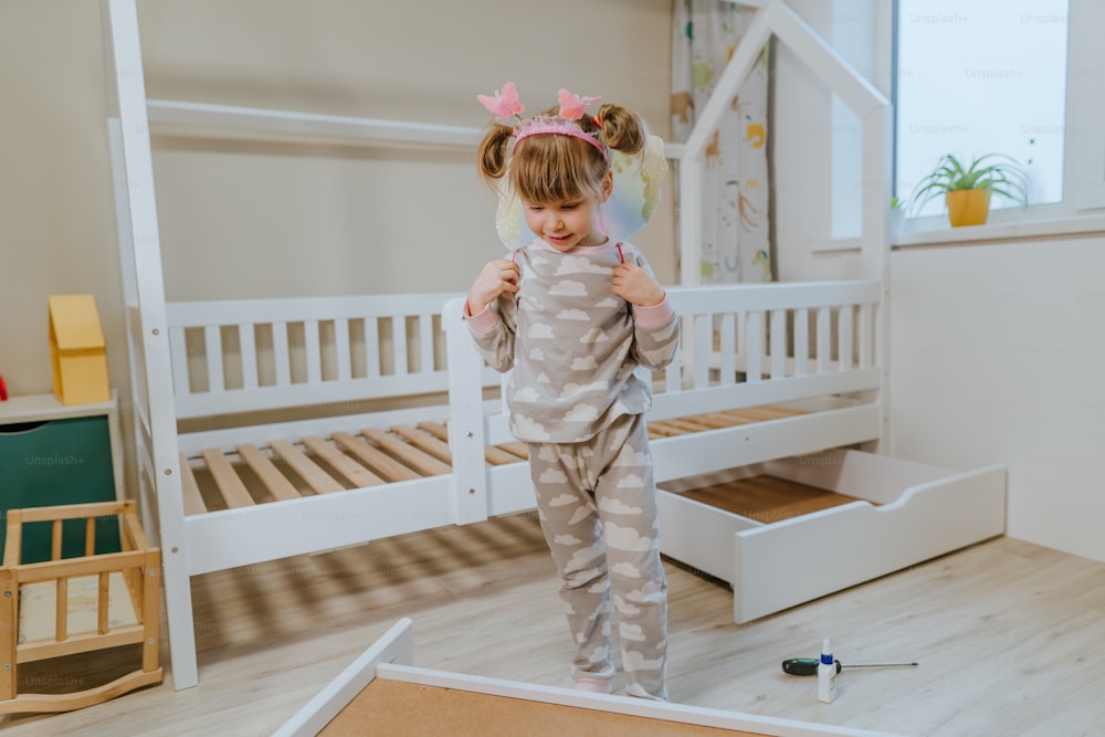 Kleines 4-jähriges Mädchen im Schlafanzug und Schmetterlingsflügel-Kostüm, das im Kinderzimmer in der Nähe des neuen Bettes spielt.