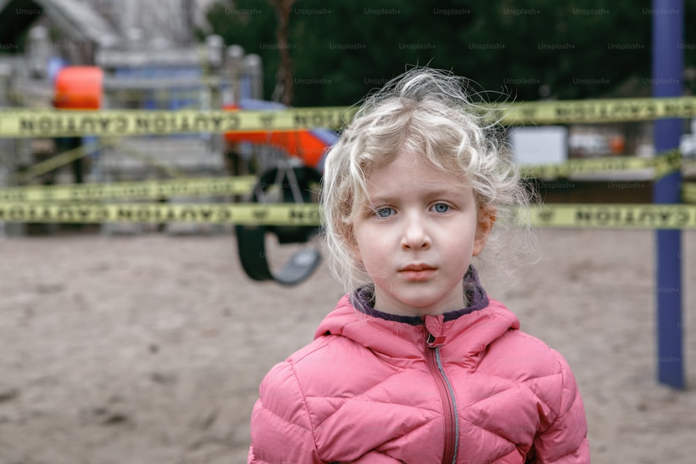 Portrait of sad upset Caucasian girl on closed playground outdoor. Kids play area locked with yellow caution tape in Toronto Canada. Coronavirus social distance quarantine.