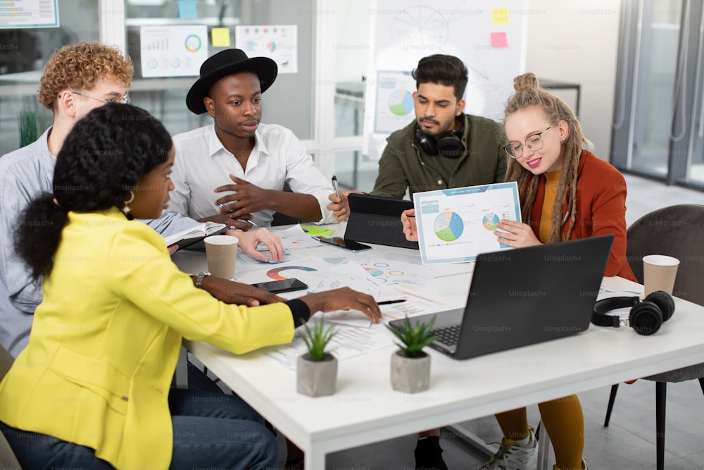 Group of young five diverse business people smiling and showing inforgaphic papers during video conference on laptop. Young hipster colleagues having working meeting online.