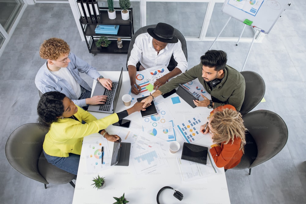 Competent IT specialists in stylish outfit working on modern gadgets while sitting together at bright office. Young multiracial people shaking hands, talking and smiling during cooperation.