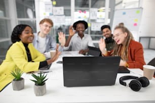 Blurred view of multiracial team of five young people having video conference on laptop at office, feeling happy and welcome their friend waving hands. Focus on laptop