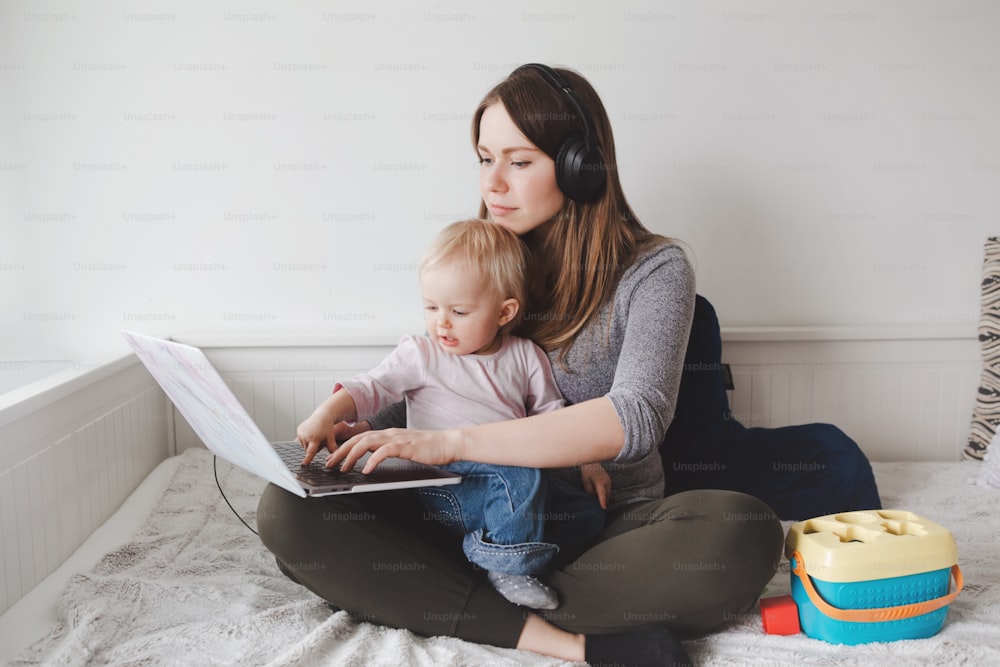Caucasian mother blogger with baby working on laptop from home. Workplace of freelance woman with kid toddler. Stay at home single mom earning money at online remote distant job.