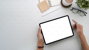 Manos de mujer sosteniendo una tableta digital de maqueta con pantalla vacía en un escritorio de oficina blanco.