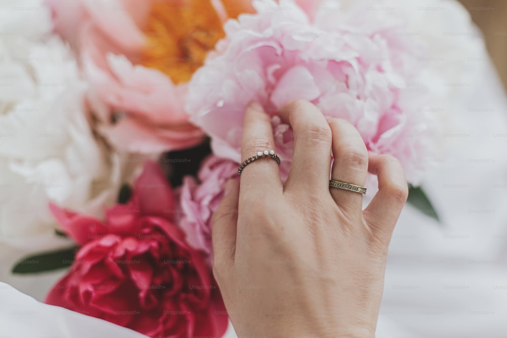 Hand mit stilvollen Ringen auf schönen Pfingstrosenstrauß. Große weiße und rosa Pfingstrosenblüten und Brauthand. Zärtliches Bild. Hochzeitsstrauß. Blumenhändler arrangiert Blumen. Feminine Essentials