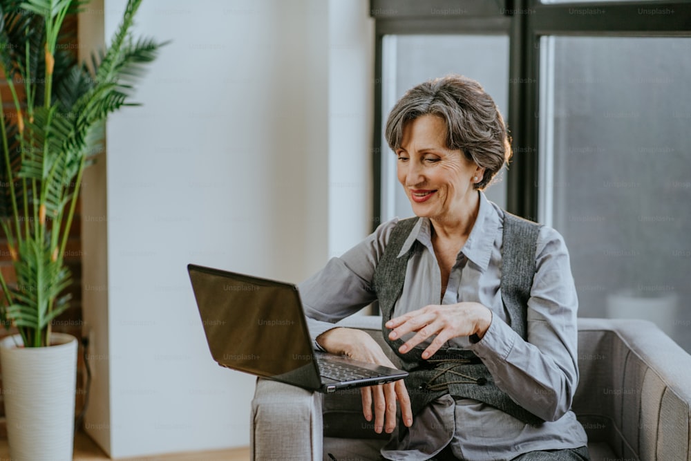 Ritratto di donna d'affari matura che utilizza il laptop per il coaching online o la consultazione seduta su una poltrona in ufficio.