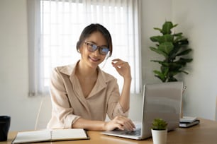 사무실에서 컴퓨터 노트북으로 일하고 카메라를 향해 웃고 있는 행복한 젊은 여성 회사원.