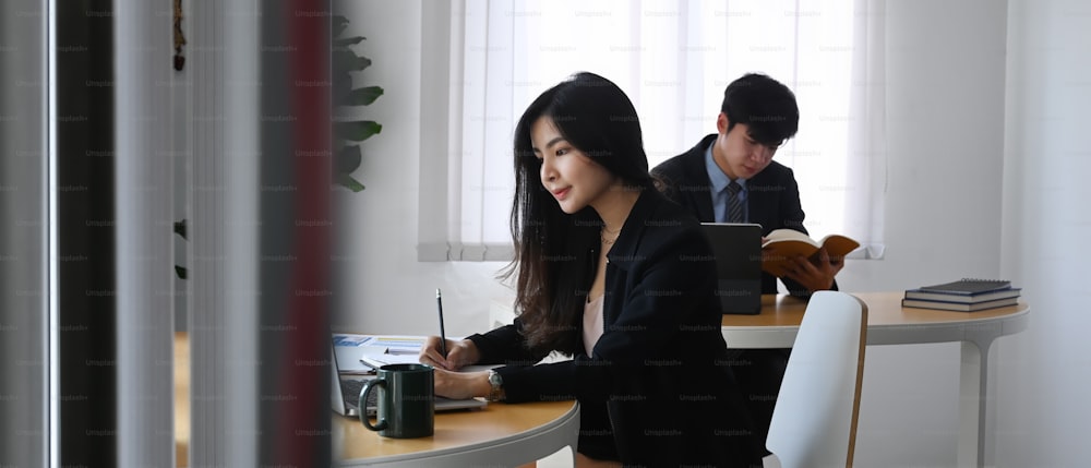若い女性実業家がノートパソコンで作業し、同僚が背景に座っている。