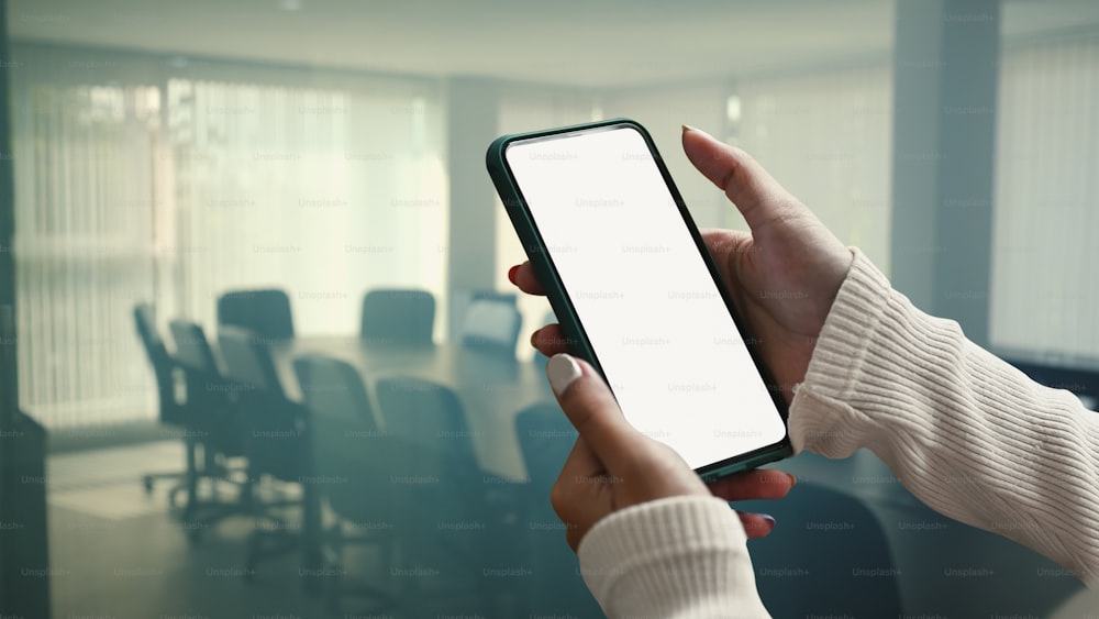Mockup image of female holding smartphone with empty screen on conference room blurred background.