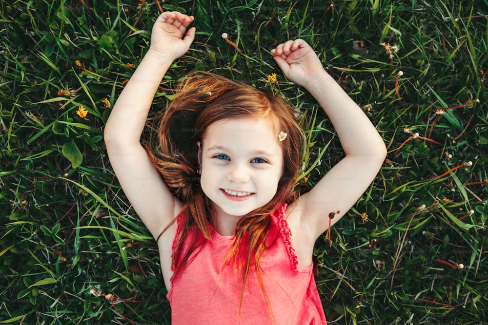 Cute smiling Caucasian girl resting in grass on meadow. Child lying on ground. Outdoor fun summer children activity. Kid having fun outside. Happy childhood lifestyle. View from top above.