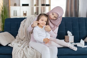 Young Muslim woman mom and her 3 years old preschool daughter child relaxing in the living room. Mom applying cream on her daughter face. Family morning care routine. Baby hygiene concept.