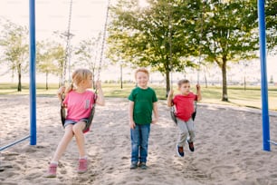 Feliz sorridente menina pré-escolar e meninos amigos balançando em balanços no playground do lado de fora no dia de verão. Conceito de estilo de vida feliz na infância. Atividade sazonal ao ar livre para crianças.