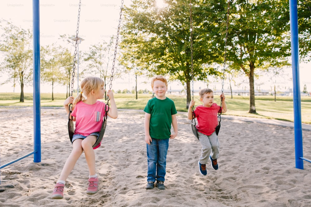 Happy smiling little preschool girl and boys friends swinging on swings at playground outside on summer day. Happy childhood lifestyle concept. Seasonal outdoor activity for kids.