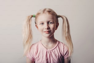 Closeup portrait of cute smiling blonde Caucasian preschool girl in pink t-shirt on light background. Child with long pig tails hair posing looking in camera. Kid expressing positive emotions.