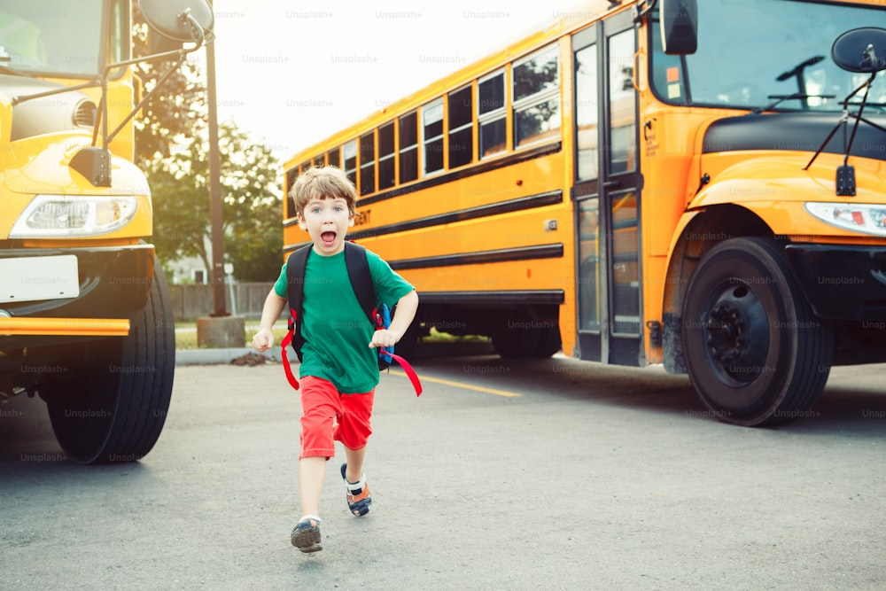 Laughing smiling Caucasian boy student kid with funny face expression walking near yellow bus on 1 September day. Education and back to school concept. Child pupil ready to learn and study.