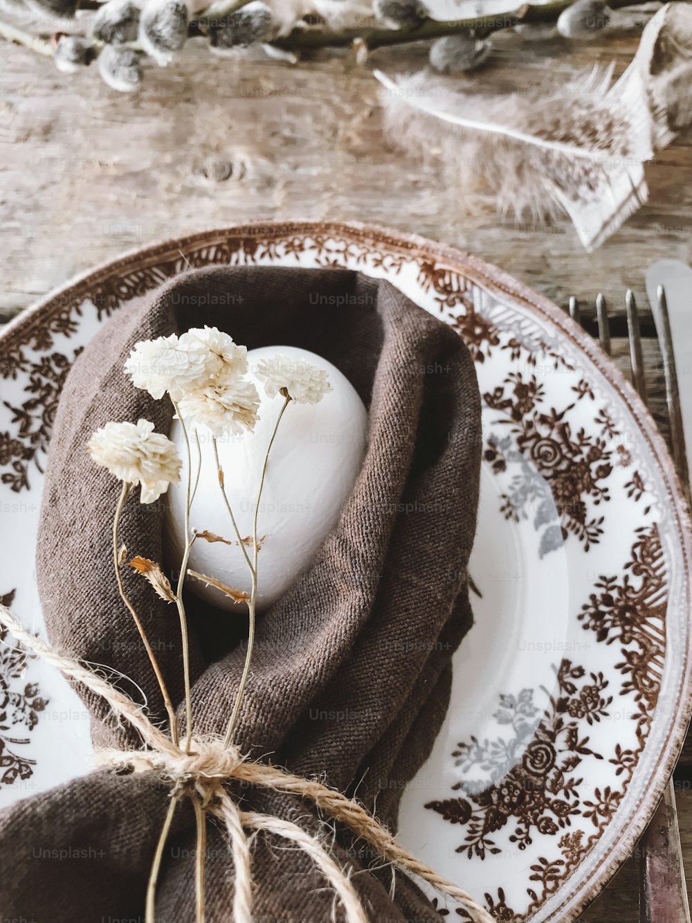 Stylish rustic Easter table setting. Natural easter egg in napkin with flowers, vintage plate and cutlery, soft feathers on aged wooden table. Rural Easter still life. Happy Easter