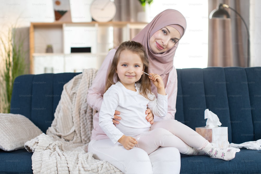 Pretty young Muslim mother and her cute little baby girl, sitting on the blue couch in modern appartment interior indoors. Mom cleans ear of her daughter with cotton swab. Close up.