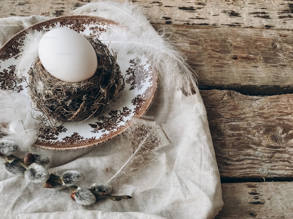 Stylish rustic Easter table setting. Natural easter egg in nest with soft feathers on vintage plate, linen napkin, pussy willow branches on aged wood. Rural Easter still life. Happy Easter