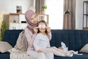 Portrait of smiling little preschool girl, sitting with her mother on the blue couch at home, having fun together, mom covering eyes of kid girl with cotton pads. Morning hygiene routine.