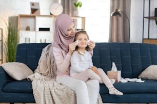 Joven madre musulmana feliz, con hiyab claro, limpiando con servilleta la cara de su linda hija de 3 años, sentada junta en el sofá azul en la acogedora sala de estar de casa.