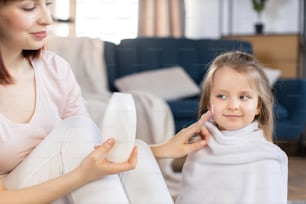 Primer plano de una joven madre sonriente aplicando loción o crema hidratante en la cara de su linda hija después de bañarse en casa. Concepto de cuidado de la piel e higiene de los niños.