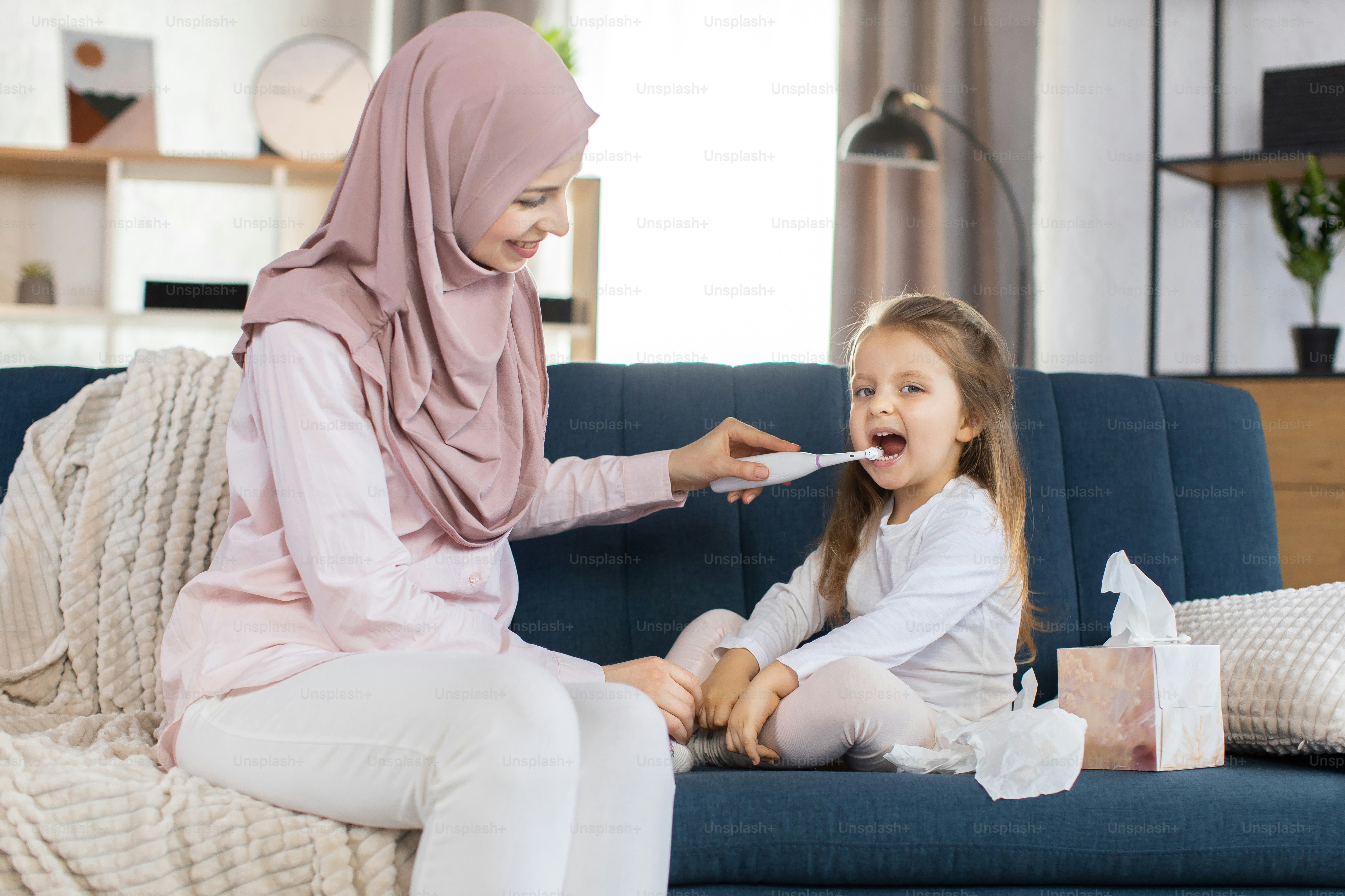 Young smiling muslim mother in hijab and her daughter, sitting at home in cozy living room, mom brushing teeth of girl with electric toothbrush. Morning hygiene routine.