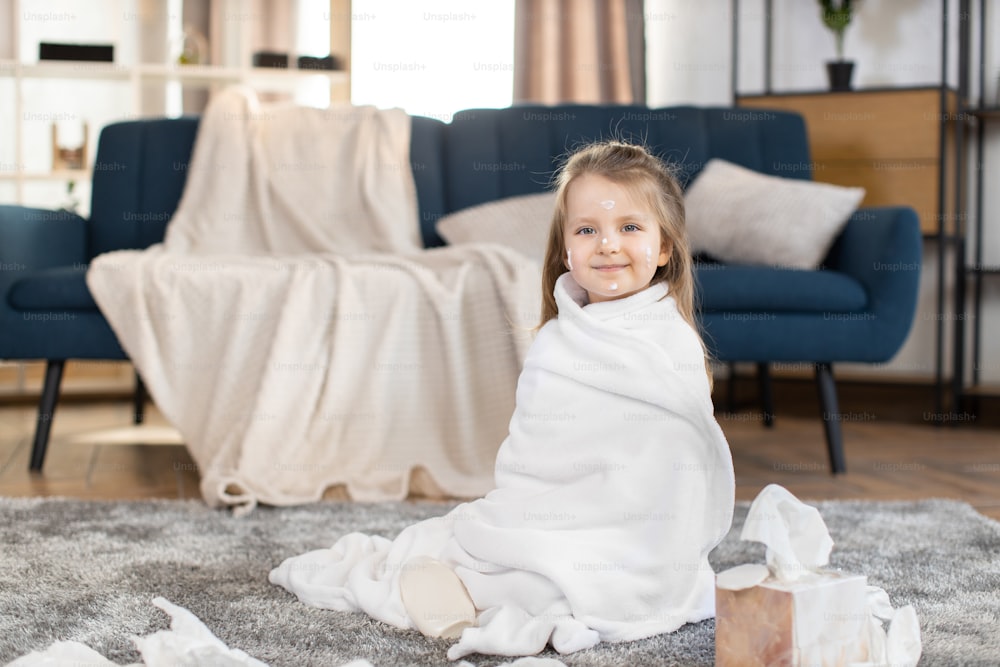 Portrait of adorable cute little 3 years old girl, sitiing on the floor at cozy room at home, wrapped in big white towel after bath, having fun with drops of body milk on face. Baby care and hygiene.