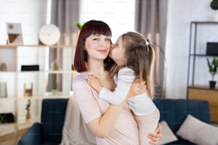 Lovely family mom and child. Happy affectionate young mother, holds her cute smiling little daughter, which is kissing mom in cheek with love, spending free time at home, posing in cozy living room.