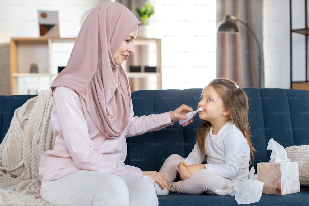 Happy arabic family, morning hygiene. Muslim mother in hijab, sitting on sofa at home, brushing teeth of her cute little daughter child with electric toothbrush, showing how to clean teeth propelry