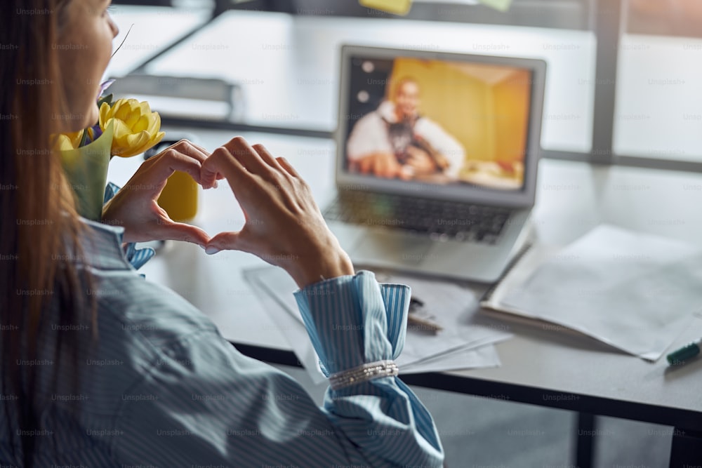 Back view of a woman with a bunch of spring flowers making a heart symbol during the video call