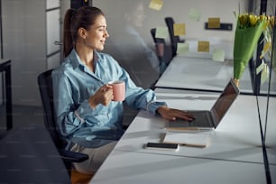 Side view of a smiling woman with a mug of coffee looking at her laptop