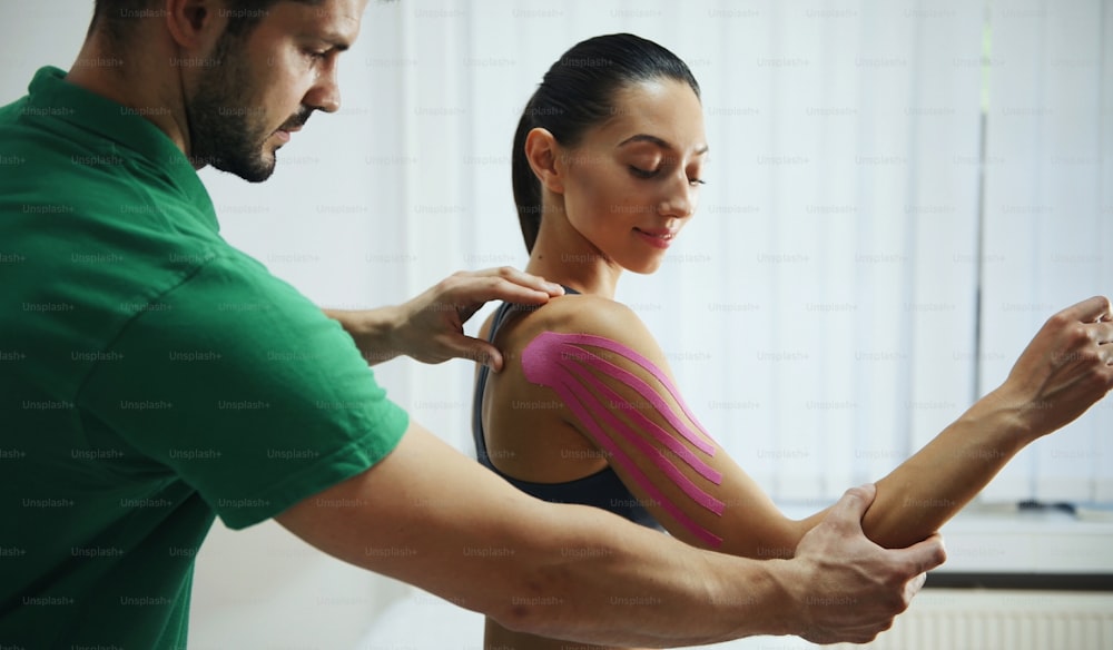 Doctor helps woman by shoulder treatment with kinesio tape.