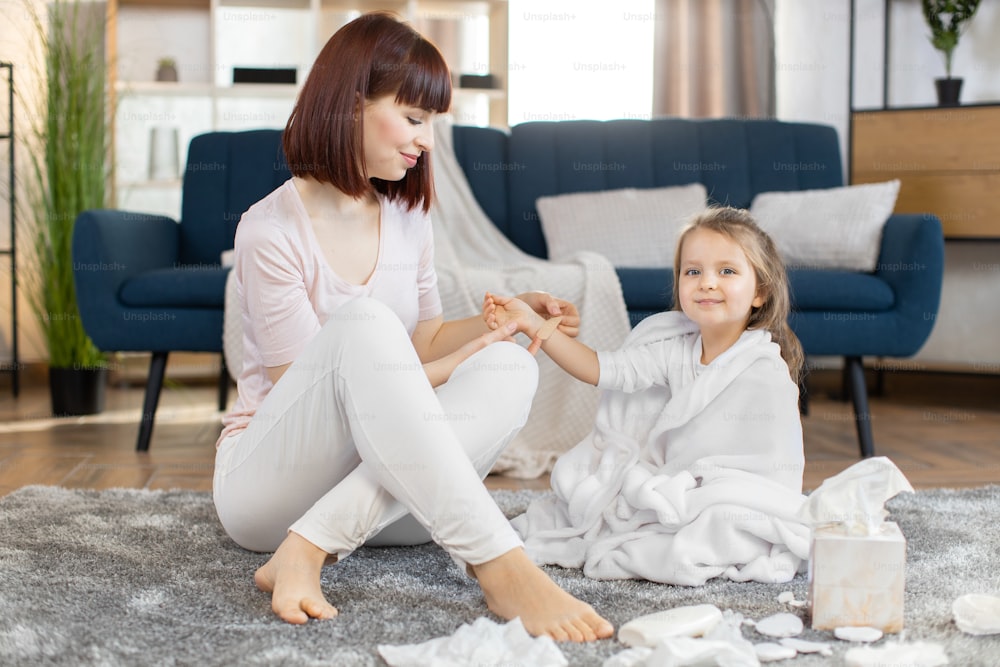 Young pretty mother applying adhesive plaster bandage on her little cute daughter arm wound, while sitting at home on the floor in living room and making care hygiene procedures.