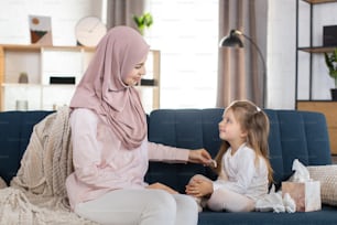 幸せな愛するアラビアの家族。ヒジャブをかぶったイスラム教徒の母親は、居心地の良いリビングルームのソファに座り、幼い娘の髪をとかしている。娘の髪型をして話す幸せな母親