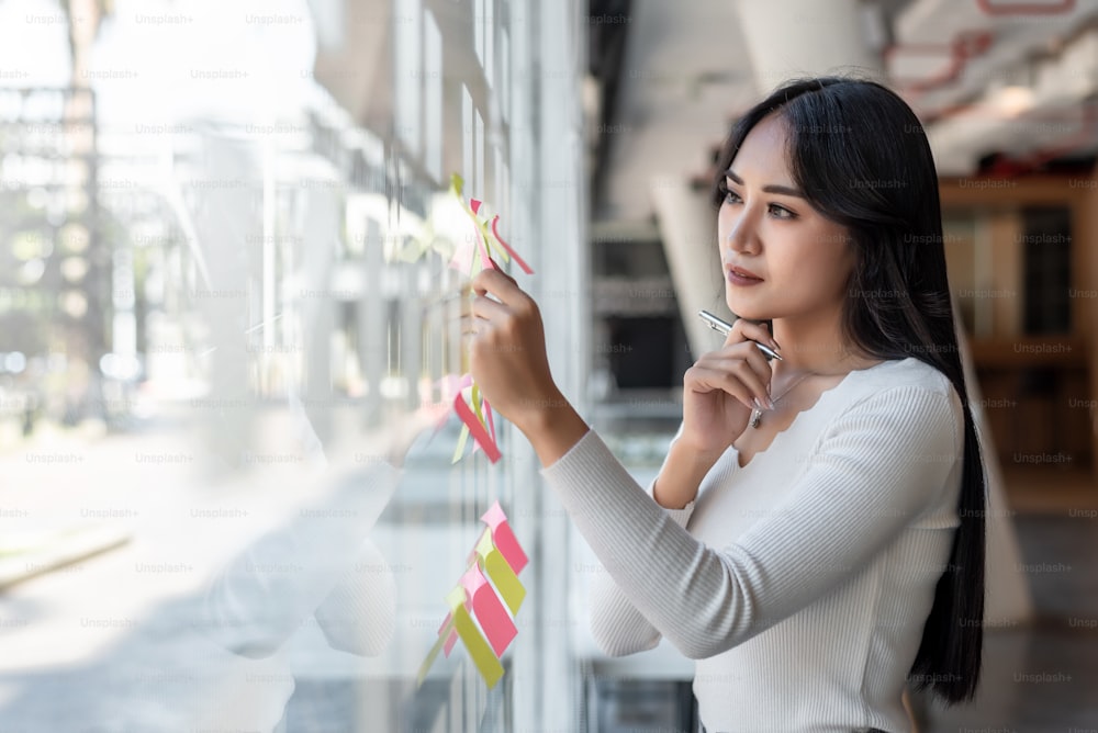 オフィスのガラス壁に付箋を貼る若いアジアの実業家の美しい魅力的な笑顔の肖像画。