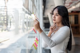 Portrait of young asian businesswoman beautiful charming smiling sticking sticky note on the glass wall in the office.