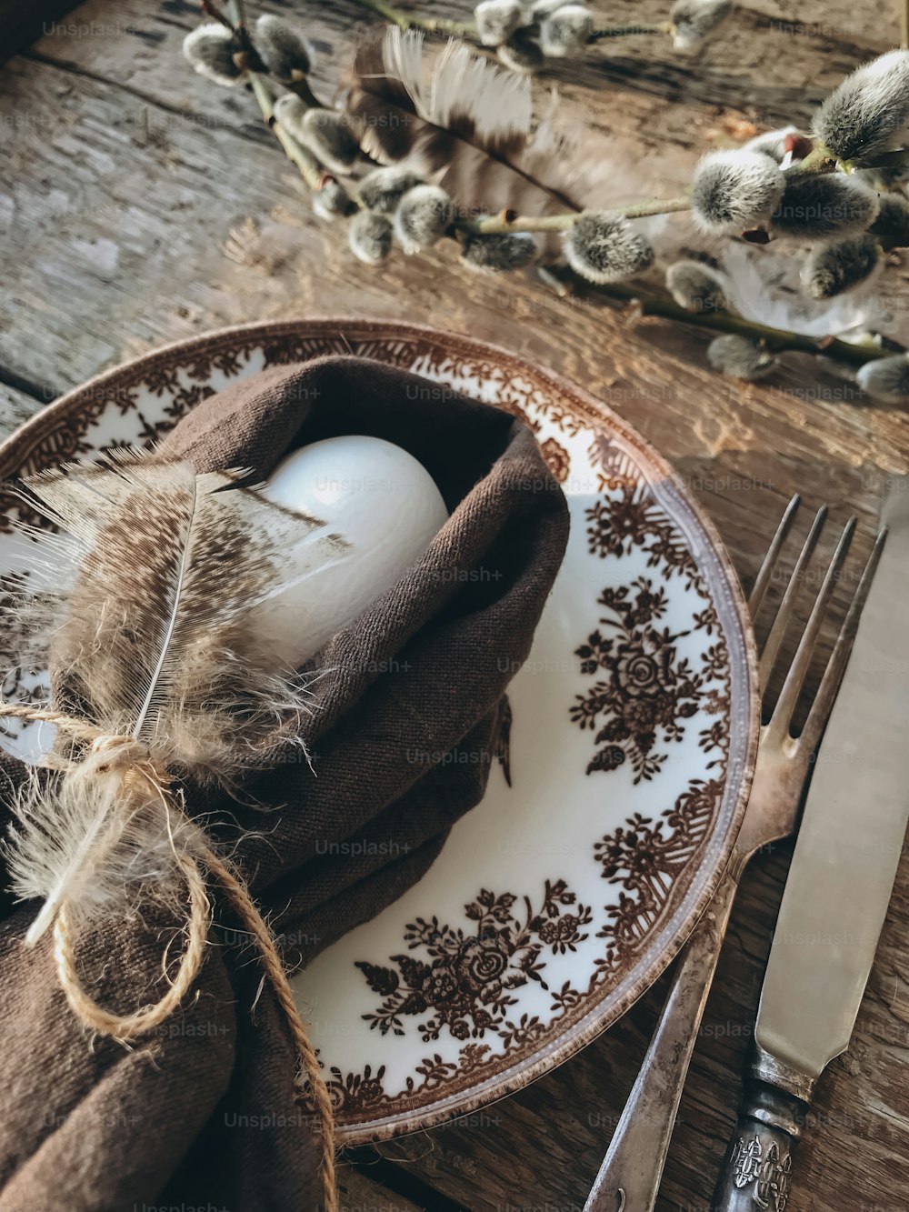 Natural easter egg in napkin with soft feathers, vintage plate and cutlery on aged wooden table. Stylish rustic Easter table setting. Rural Easter still life. Happy Easter