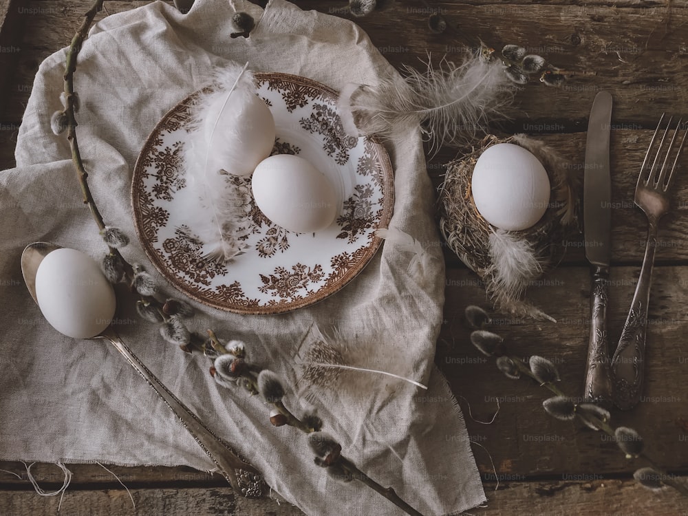 Natural easter eggs, feathers, vintage plate and cutlery, napkin, pussy willow branches on aged wooden table. Rural Easter still life. Happy Easter. Stylish rustic Easter table setting flat lay.