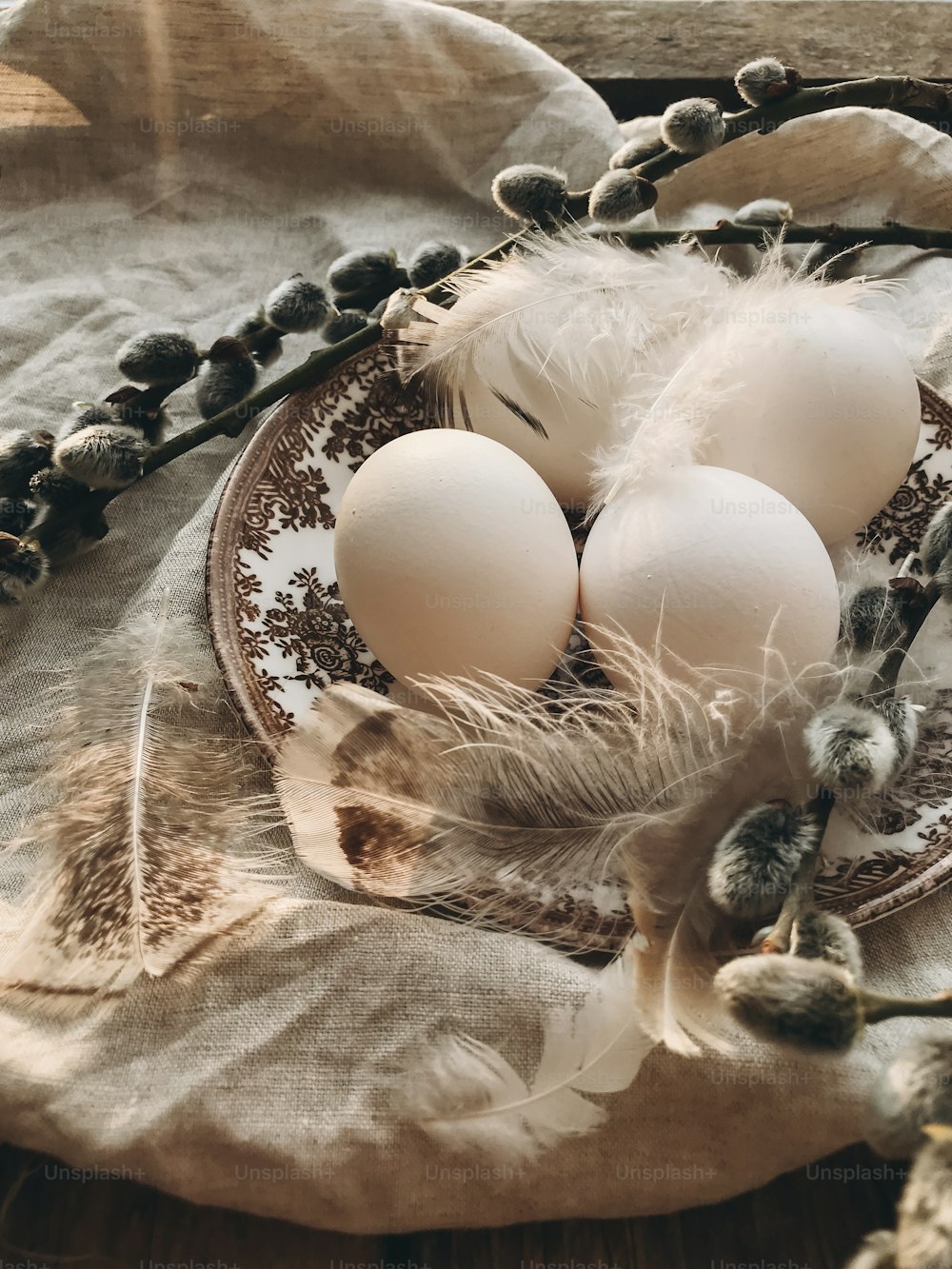 Rural Easter still life. Natural Easter eggs on vintage plate, feathers, pussy willow branches on napkin on aged wooden table in sunny light.  Happy Easter. Stylish rustic table setting