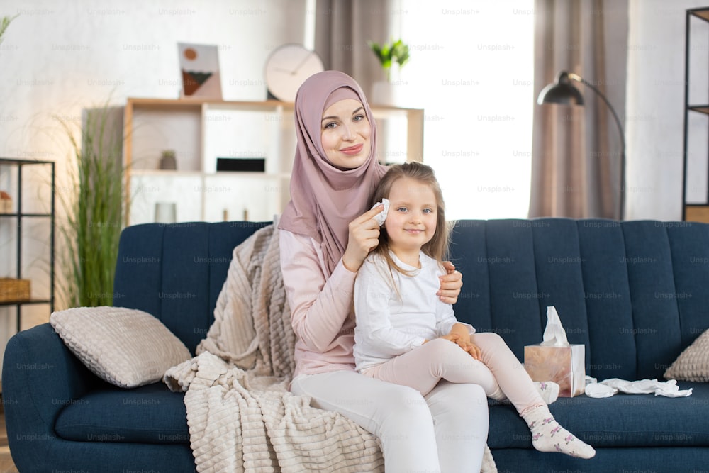 Young Arabian mom in headscarf holds her little pretty girl child on her knees, sitting on the sofa in living room at home. Smiling mother wiping baby s face with paper napkin.