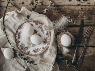 Rural Easter still life. Natural easter eggs, feathers, vintage plate and cutlery, napkin, pussy willow branches on aged wooden table, flat lay. Happy Easter. Stylish rustic Easter table setting