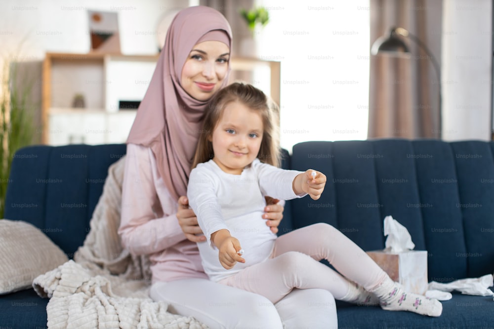 Young smiling Arabian mother with her cute little daughter, sitting on the blue sofa in cozy living room. Pretty child showing hygienic cotton swabs to camera. Hygiene and care concept.