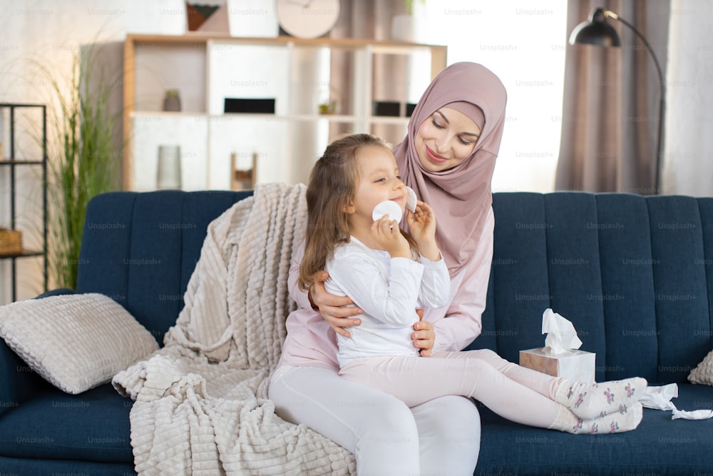 Adorabile mamma musulmana premurosa, seduta sul divano blu di casa e tenendo la sua bambina sulle ginocchia. Il simpatico bambino felice fa procedure igieniche e si pulisce il viso con dischetti di cotone.