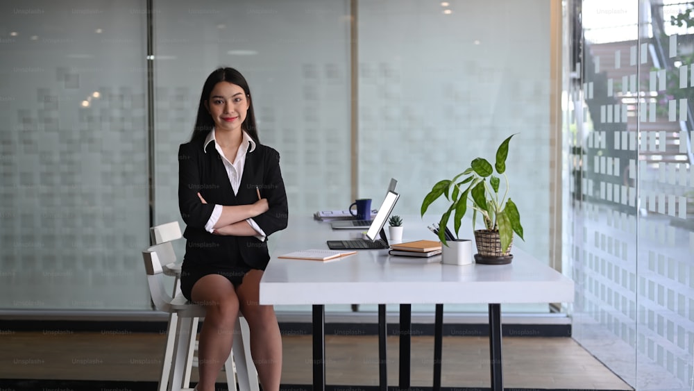 Confident businesswoman sin black suit sitting with arms crossed in modern office.
